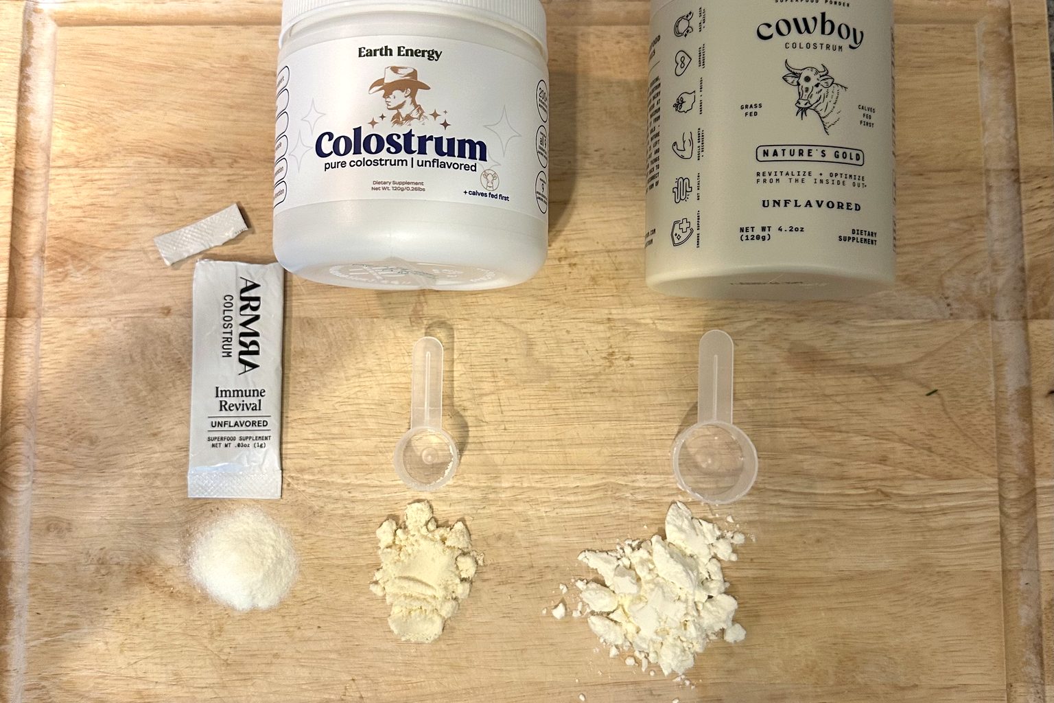 Overhead photo of ARMRA's 1 gram packet, Earth Energy's 2 gram scoop and Cowboy Colostrum's 3 gram scoop of powder side by side on a cutting board